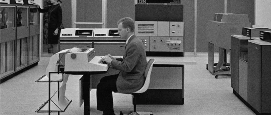 Man typing at a computer terminal in a mid-century machine room, surrounded by mainframe equipment